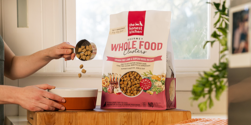One bag of The Honest Kitchen clusters dry dog food, with a bowl sitting on a kitchen counter, hands pouring food into bowl