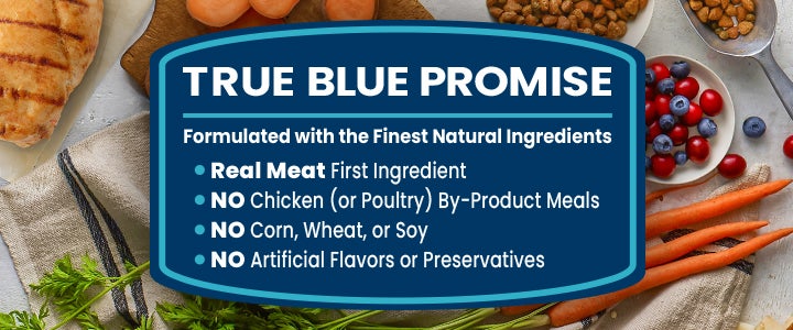 A marketing banner with a dark blue graphic badge centered over a background of fresh food ingredients. The text inside the badge reads "TRUE BLUE PROMISE," followed by "Formulated with the Finest Natural Ingredients." It lists four bullet points: "Real Meat First Ingredient," "NO Chicken (or Poultry) By-Product Meals," "NO Corn, Wheat, or Soy," and "NO Artificial Flavors or Preservatives." The background features grilled chicken breast, fresh carrots, blueberries, cranberries, kibble, and fresh parsley on a rustic cloth surface.